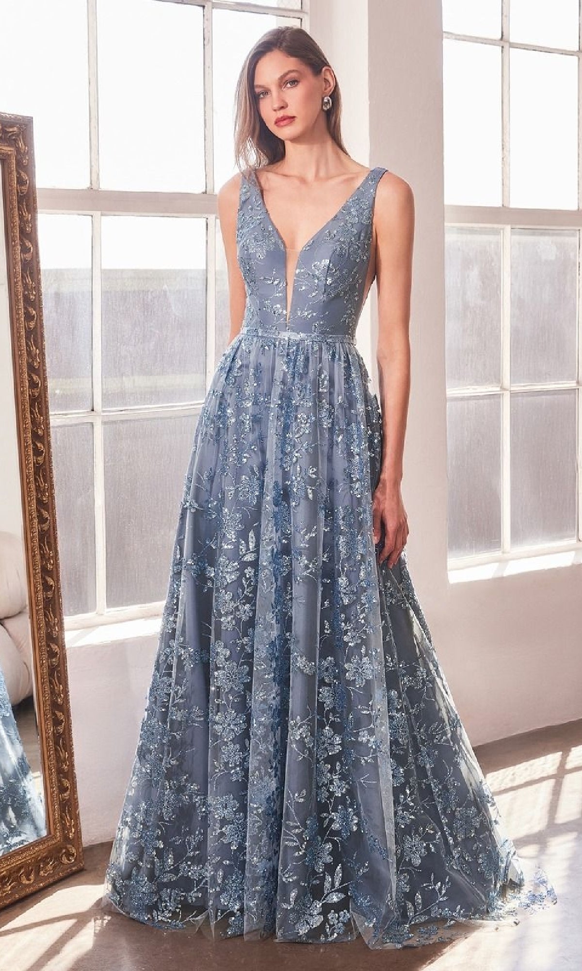 A woman stands indoors by a window and mirror wearing the Amoris T606, an embroidered-lace long prom dress featuring a sleeveless, deep V-neck, floor-length blue design with floral details.